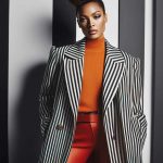 A woman stands confidently against a background of vertical black and white stripes. She is wearing a stylish outfit consisting of an orange turtleneck sweater, red pants, and a black and white striped trench coat. The blazer has wide lapels and gold buttons. Her hair is styled in an elegant updo, and she has a poised expression. The combination of her outfit and the background creates a striking visual contrast.