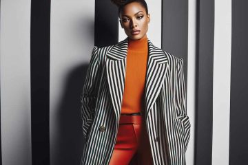 A woman stands confidently against a background of vertical black and white stripes. She is wearing a stylish outfit consisting of an orange turtleneck sweater, red pants, and a black and white striped trench coat. The blazer has wide lapels and gold buttons. Her hair is styled in an elegant updo, and she has a poised expression. The combination of her outfit and the background creates a striking visual contrast.