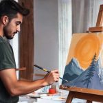 A man with short dark hair and a beard is painting on a canvas set on an easel in a well-lit studio. He is wearing a dark green t-shirt and is focused on his work, holding a paintbrush with his right hand. The painting depicts a landscape with mountains, a tall evergreen tree in the foreground, and a large orange sun in a bright sky. The room has large windows with light streaming in, casting a soft glow, and there are various art supplies and tools on a table in the background.