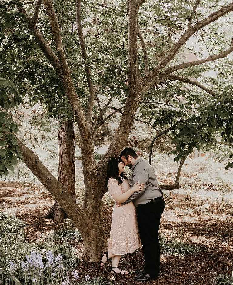 The image features a couple standing closely together under a large tree in a natural, wooded setting. The woman, wearing a light pink dress and black sandals, has her arms around the man's shoulders. The man, dressed in a gray shirt and black pants, leans slightly forward with his arms around her waist. Their foreheads touch as they share an intimate moment. The ground is covered with mulch and small plants, including a cluster of purple flowers in the foreground. The scene is softly lit, creating a serene and romantic atmosphere.