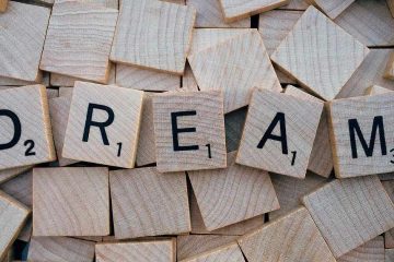 The image shows wooden Scrabble-style tiles scattered in a pile. Five tiles in the center spell out the word "DREAM" in black, uppercase letters. Each tile has a small number in the lower right corner indicating its point value, such as "D" worth 2 points and "M" worth 3 points. The surrounding tiles are blank or face down, creating a textured wooden background.