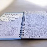 A spiral-bound notebook is open on a light-colored surface, with two pages filled with handwritten text and doodles. The left page reads, "Messy doodles and imperfections are allowed in here. That’s the purpose of this book." Surrounding the text are swirly patterns and abstract designs, including curved lines and shapes. The right page is filled with various intricate doodles, including wavy patterns, spirals, and abstract shapes, along with small handwritten notes. The notebook and its creative contents appear to be illuminated by soft natural light.