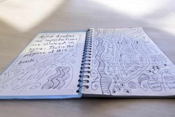 A spiral-bound notebook is open on a light-colored surface, with two pages filled with handwritten text and doodles. The left page reads, "Messy doodles and imperfections are allowed in here. That’s the purpose of this book." Surrounding the text are swirly patterns and abstract designs, including curved lines and shapes. The right page is filled with various intricate doodles, including wavy patterns, spirals, and abstract shapes, along with small handwritten notes. The notebook and its creative contents appear to be illuminated by soft natural light.