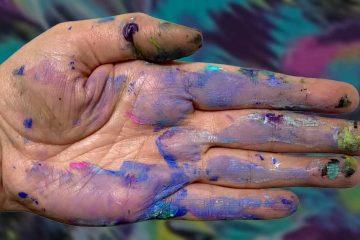 A close-up of an open human hand covered in smudges and smears of various paint colors, including purple, blue, pink, green, yellow, and black. The background is blurred but shows similarly colorful, abstract patterns, suggesting an artistic or creative environment.