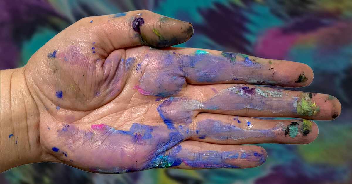 A close-up of an open human hand covered in smudges and smears of various paint colors, including purple, blue, pink, green, yellow, and black. The background is blurred but shows similarly colorful, abstract patterns, suggesting an artistic or creative environment.