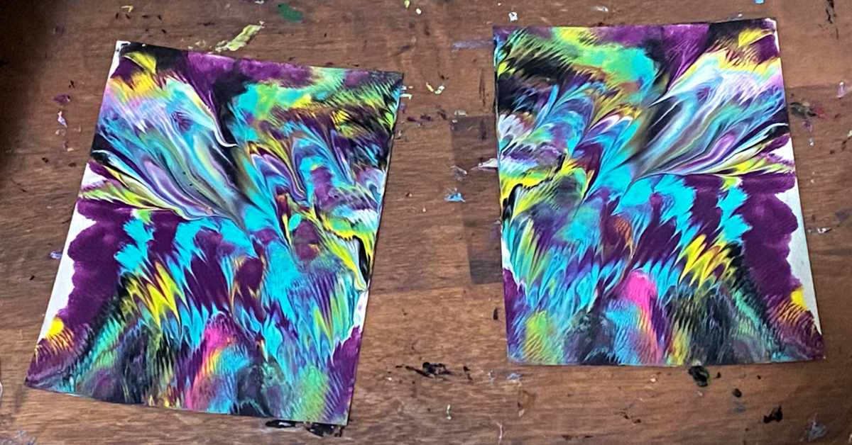Two sheets of paper with vibrant abstract paintings lie on a wooden surface. The artwork features swirling patterns in bold colors including teal, yellow, magenta, black, white, and purple, creating a marbled or fluid effect. The designs on both sheets are similar but not identical, suggesting a mirrored or monoprint technique. Paint smudges are visible on the wooden surface beneath.