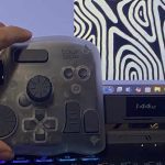 A hand is holding a TourBox controller in front of a computer monitor. The controller is semi-transparent with multiple dials, buttons, and knobs designed for creative software use. On the monitor, there is abstract black-and-white artwork with organic, contour-like patterns on the right side, and part of a digital art program interface is visible on the left. The monitor displays a 144Hz refresh rate label and various app icons on the taskbar.