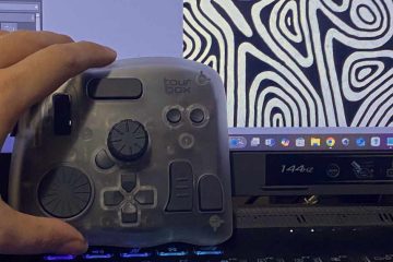 A hand is holding a TourBox controller in front of a computer monitor. The controller is semi-transparent with multiple dials, buttons, and knobs designed for creative software use. On the monitor, there is abstract black-and-white artwork with organic, contour-like patterns on the right side, and part of a digital art program interface is visible on the left. The monitor displays a 144Hz refresh rate label and various app icons on the taskbar.