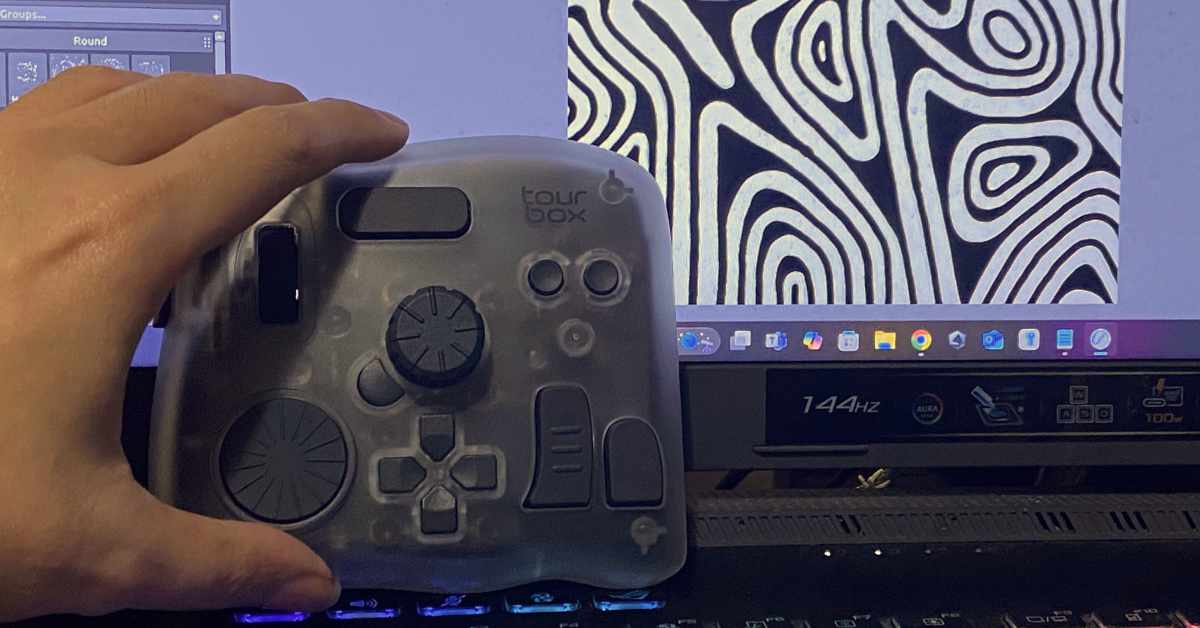 A hand is holding a TourBox controller in front of a computer monitor. The controller is semi-transparent with multiple dials, buttons, and knobs designed for creative software use. On the monitor, there is abstract black-and-white artwork with organic, contour-like patterns on the right side, and part of a digital art program interface is visible on the left. The monitor displays a 144Hz refresh rate label and various app icons on the taskbar.