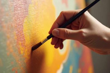 Close-up of a hand holding a paintbrush, applying warm-toned paint—mainly shades of yellow and orange—onto a textured canvas. The painting surface shows blended areas of color, suggesting an abstract or impressionistic style. The lighting highlights the brushstrokes and texture of the canvas.