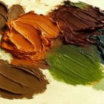 A close-up photo of thick, textured blobs of oil paint in earthy and autumn-inspired tones arranged on a light-colored palette. The colors closely resemble the October 2025 palette, including dark brown, burnt orange, burgundy, beige, green, and blue-gray hues, with visible brush strokes adding texture and depth.