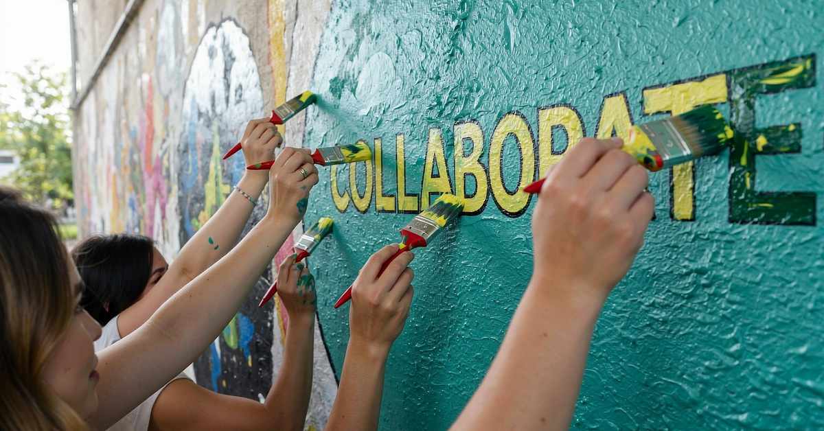 Several people are shown painting a mural on a textured wall. Multiple hands holding paintbrushes are working together to paint the word "COLLABORATE" in bold, yellow and green block letters against a teal background. The image emphasizes teamwork and community involvement in creating public art.