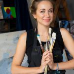 A woman wearing a black apron stands in an art studio, holding a bundle of paintbrushes in both hands. She has light brown hair tied up and is smiling slightly. Behind her are several abstract and colorful paintings, an easel, a wooden ladder, and shelving with more artwork. The setting appears to be a creative workspace for painting.