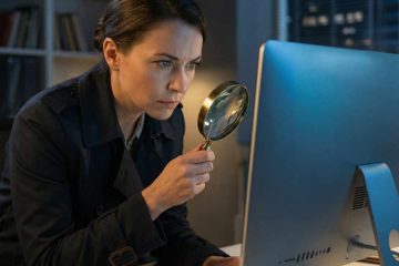 A woman sits at a desk in a dimly lit office, closely examining a large desktop computer monitor with a handheld magnifying glass. She wears a dark jacket and leans forward with a focused expression. A desk lamp illuminates the workspace, and shelves. Windows with city lights are visible in the background.