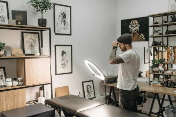 Interior of a tattoo studio with white walls covered in framed black-and-white artwork. A tattoo artist wearing a beanie and white T-shirt stands near a lit ring light, adjusting his hat. In the foreground is a black tattoo chair. Wooden shelves display plants, jars, and art prints, while a desk along the wall holds equipment and decorative items.