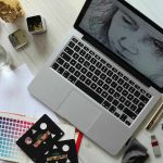 Overhead view of a creative workspace with a laptop displaying a black-and-white face sketch, surrounded by drawing tools, color swatches, pencils, a glass of water, and paper sketches on a light wooden desk.