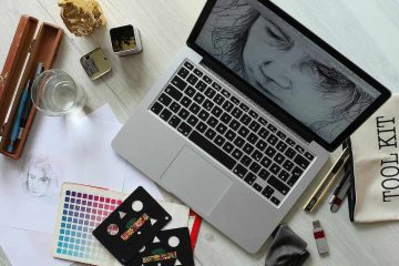 Overhead view of a creative workspace with a laptop displaying a black-and-white face sketch, surrounded by drawing tools, color swatches, pencils, a glass of water, and paper sketches on a light wooden desk.