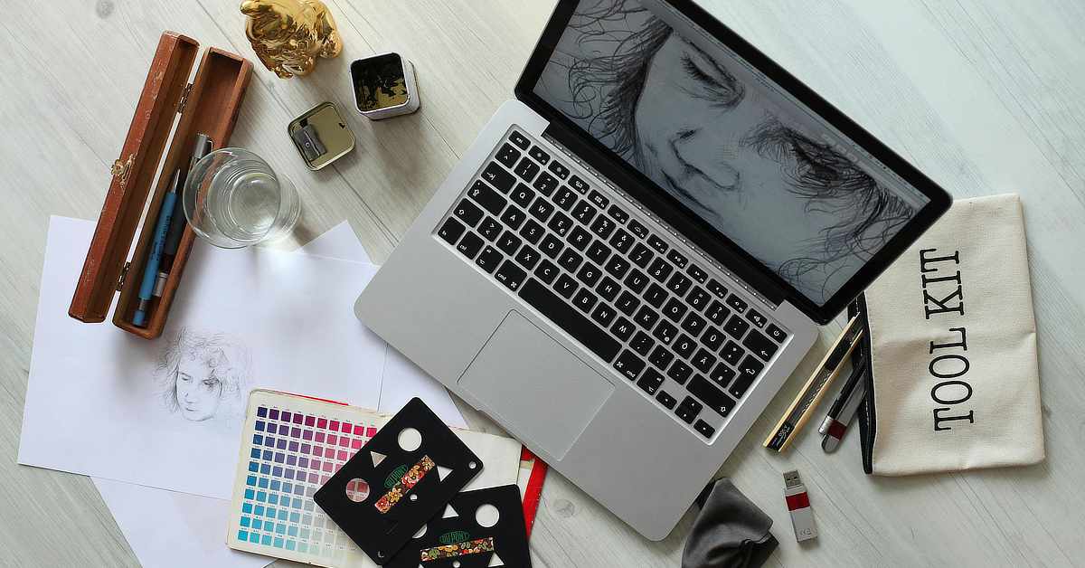 Overhead view of a creative workspace with a laptop displaying a black-and-white face sketch, surrounded by drawing tools, color swatches, pencils, a glass of water, and paper sketches on a light wooden desk.