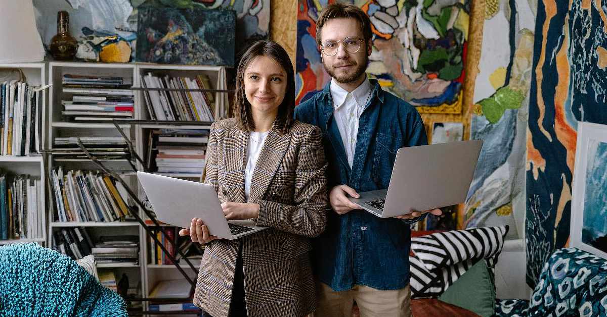 Two people standing side by side in an art studio, each holding a laptop, with shelves of books and colorful abstract paintings in the background.