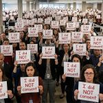 A large group of people standing in an open-plan office, each holding a printed sign that reads “I did NOT consent.” The crowd fills the space from foreground to background, with desks and computer monitors visible between rows. The individuals face forward toward the camera, holding their signs at chest level, creating a repeated pattern of the same message across the entire image.