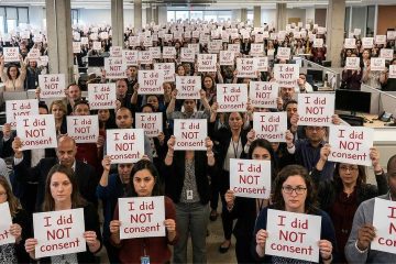 A large group of people standing in an open-plan office, each holding a printed sign that reads “I did NOT consent.” The crowd fills the space from foreground to background, with desks and computer monitors visible between rows. The individuals face forward toward the camera, holding their signs at chest level, creating a repeated pattern of the same message across the entire image.