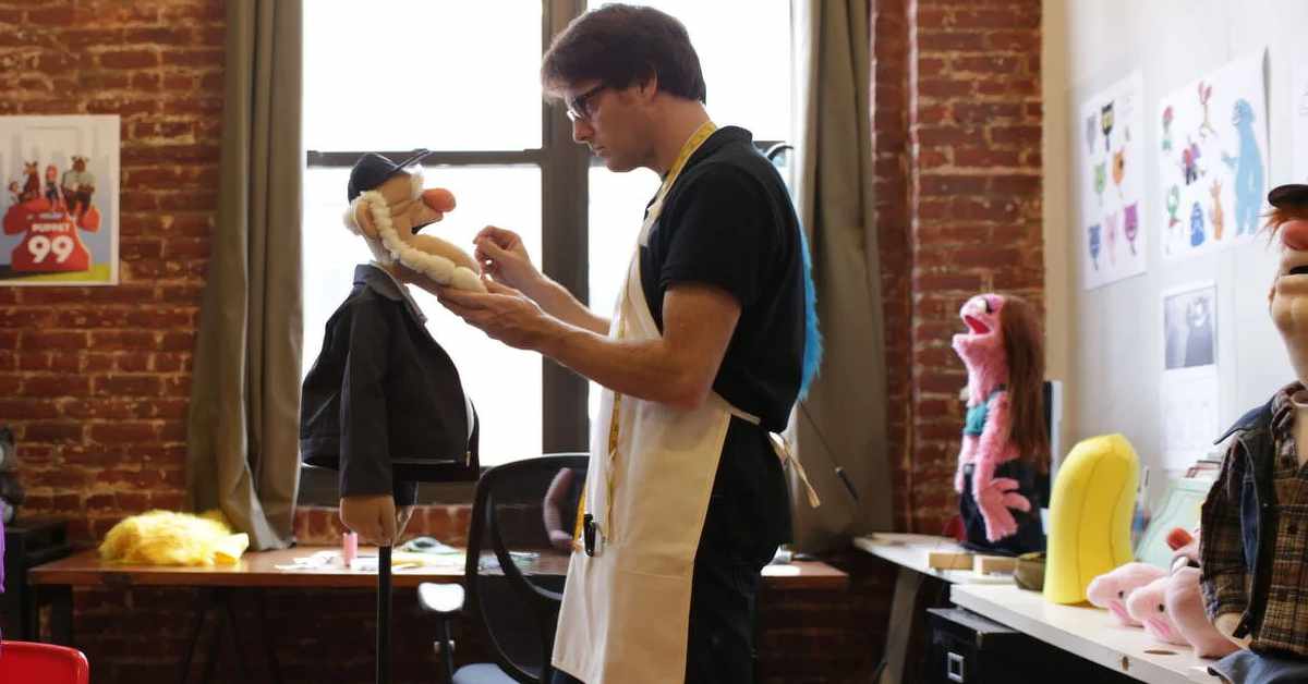 A person wearing a measuring tape around their neck adjusts the mouth of a puppet mounted on a stand in a workshop. The room has exposed brick walls, tall windows, sewing and crafting tables, and several colorful puppets and puppet parts visible in the background, including a pink puppet, a yellow foam piece, and another partially visible puppet at the right edge.