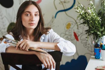A person with long brown hair wearing a white button-up shirt leans on the back of a dark wooden chair while holding several paintbrushes. They face the camera with a neutral expression. In the background, a wall features abstract painted shapes and lines. To the right, a white desk holds cups filled with colorful markers and pens, a watering can, scattered art supplies, and a vase of white flowers near a window.