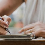 Close-up of a person writing in a notebook with a blue pen while seated at a table, with only their hands and part of their torso visible.