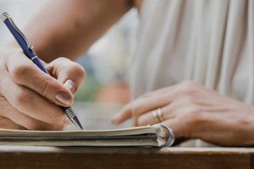 Close-up of a person writing in a notebook with a blue pen while seated at a table, with only their hands and part of their torso visible.