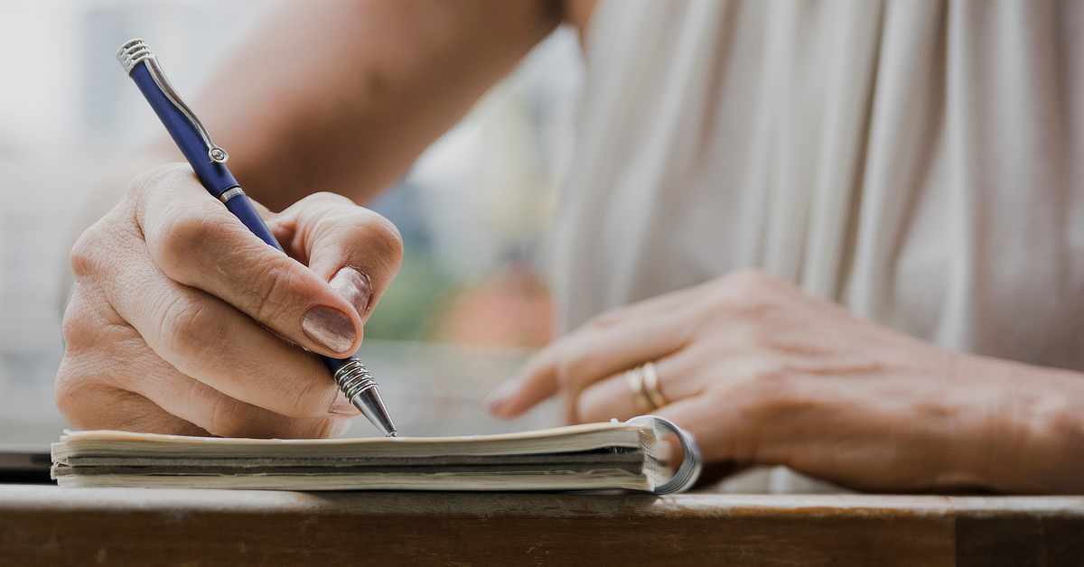 Close-up of a person writing in a notebook with a blue pen while seated at a table, with only their hands and part of their torso visible.